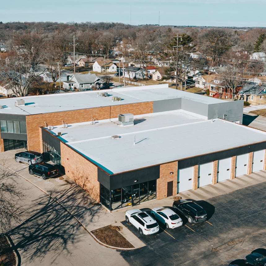 Aerial view of a commercial building with a white flat roof and brick exterior, surrounded by parked cars and residential houses in the background.