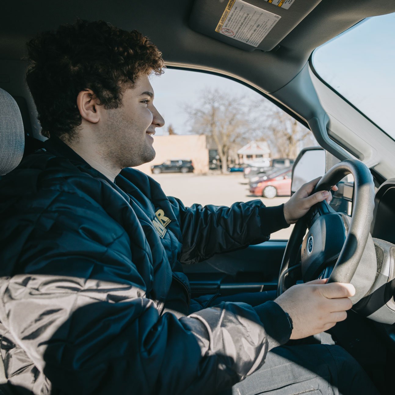 Smiling young man with curly hair wearing a black jacket driving a car during daytime.