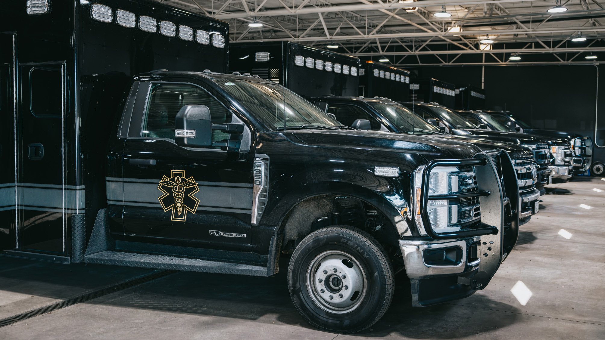 Row of black emergency medical service trucks parked indoors with visible medical emblem on the door of the closest truck.