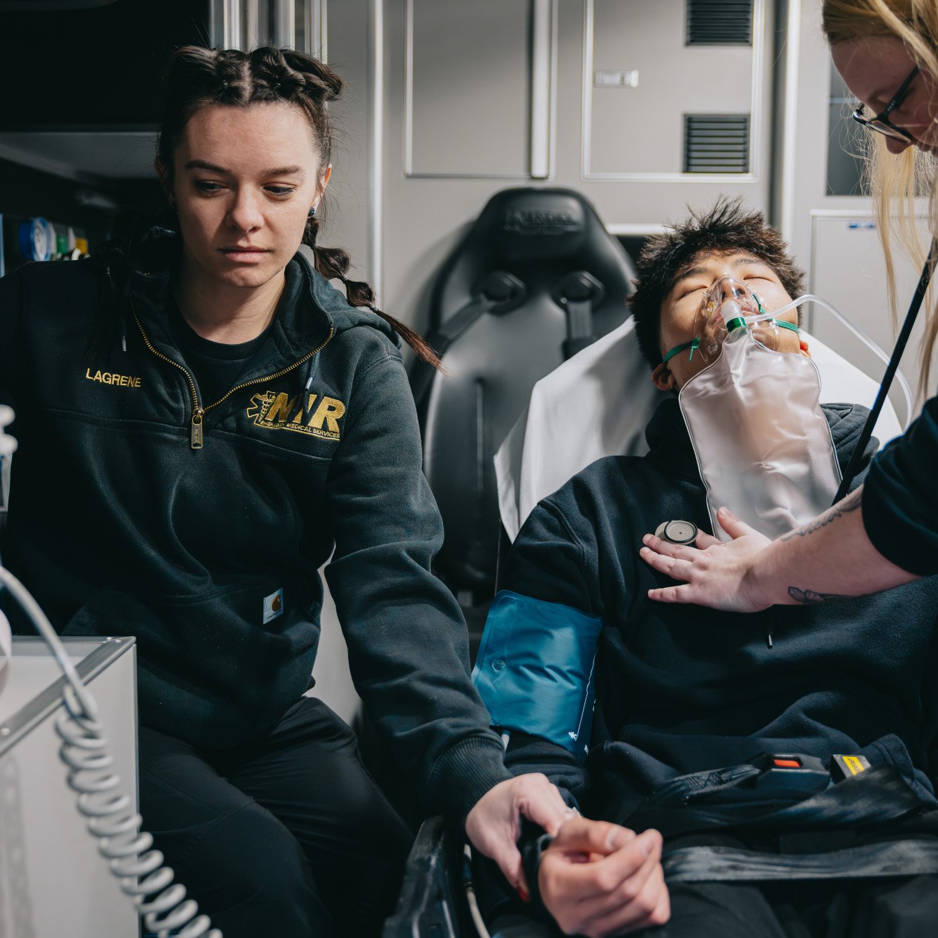 Paramedics attending to a patient lying on a stretcher inside an ambulance, with one checking the patient's chest and another holding the patient's hand.