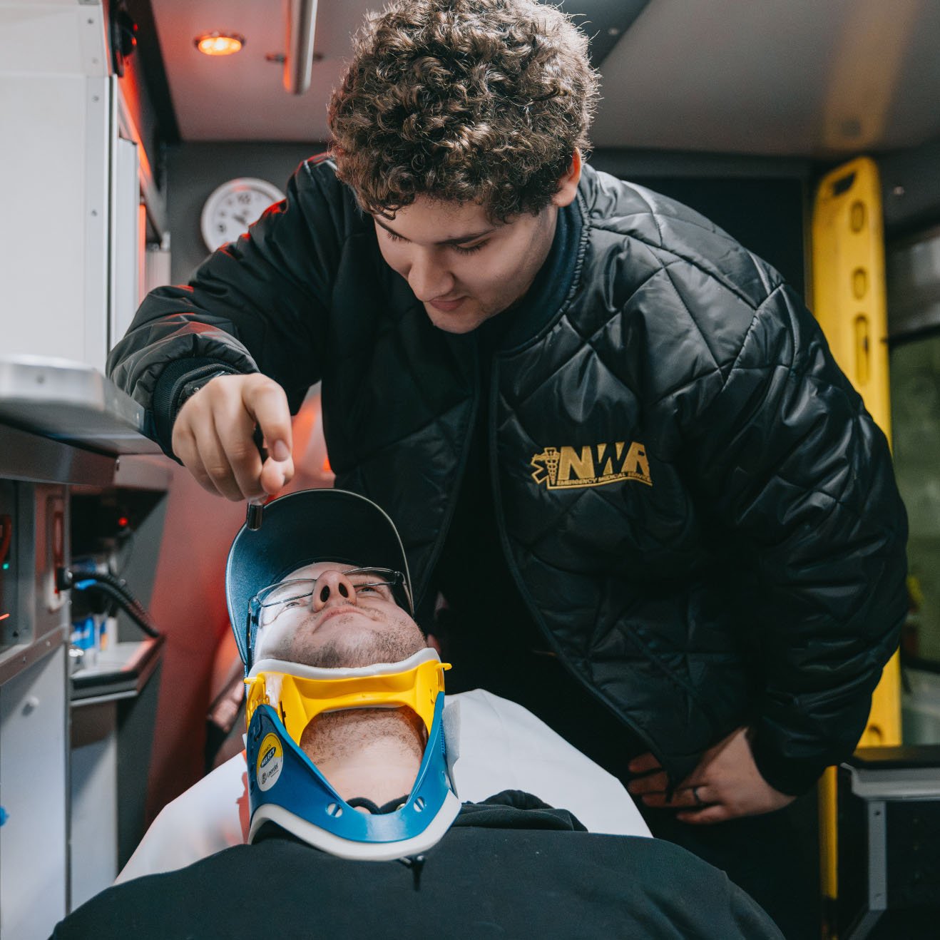 Emergency medical technician wearing a jacket with NWA logo attending to a man lying on a stretcher with a cervical collar and glasses inside an ambulance.