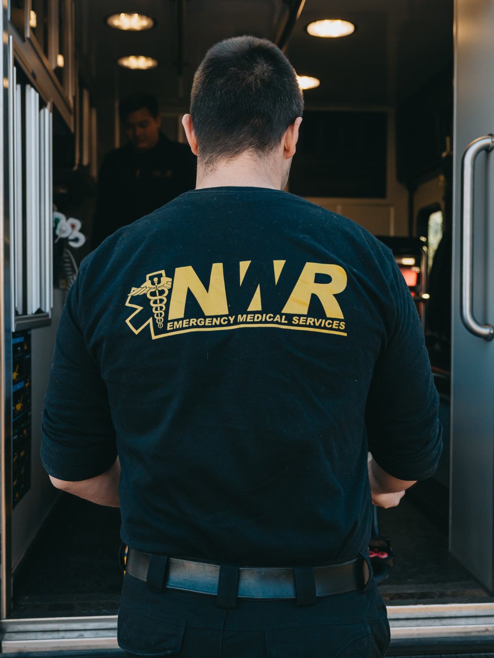 Person wearing a black NWR Emergency Medical Services shirt, standing at the entrance of an ambulance.