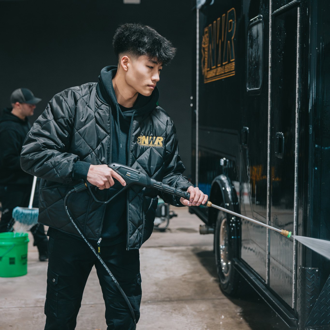 Young man in a black NWR jacket pressure washing a black vehicle while another person cleans in the background.
