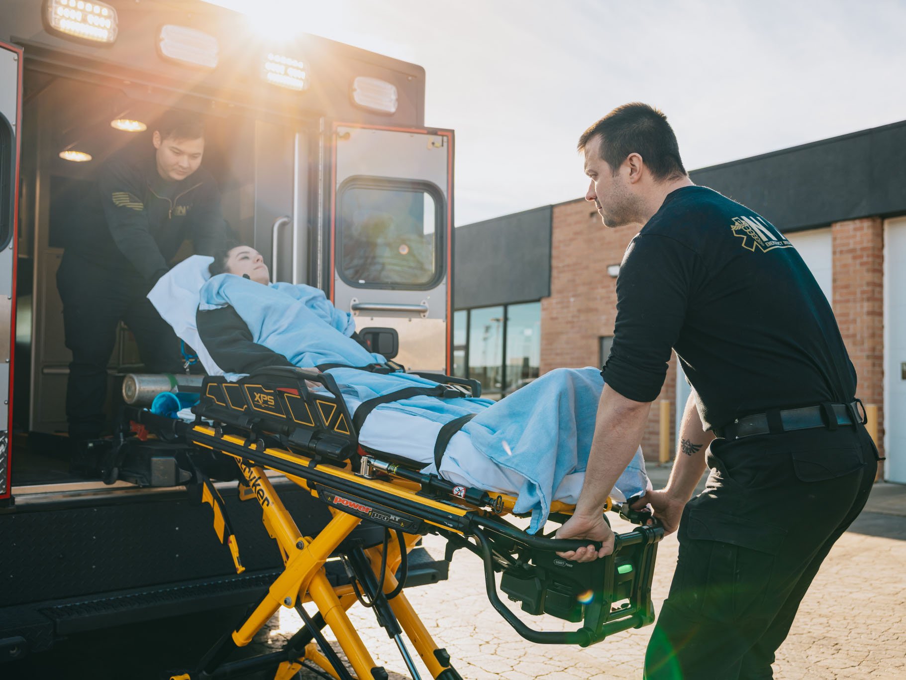 Two paramedics loading a woman covered with a blue blanket on a stretcher into an ambulance.