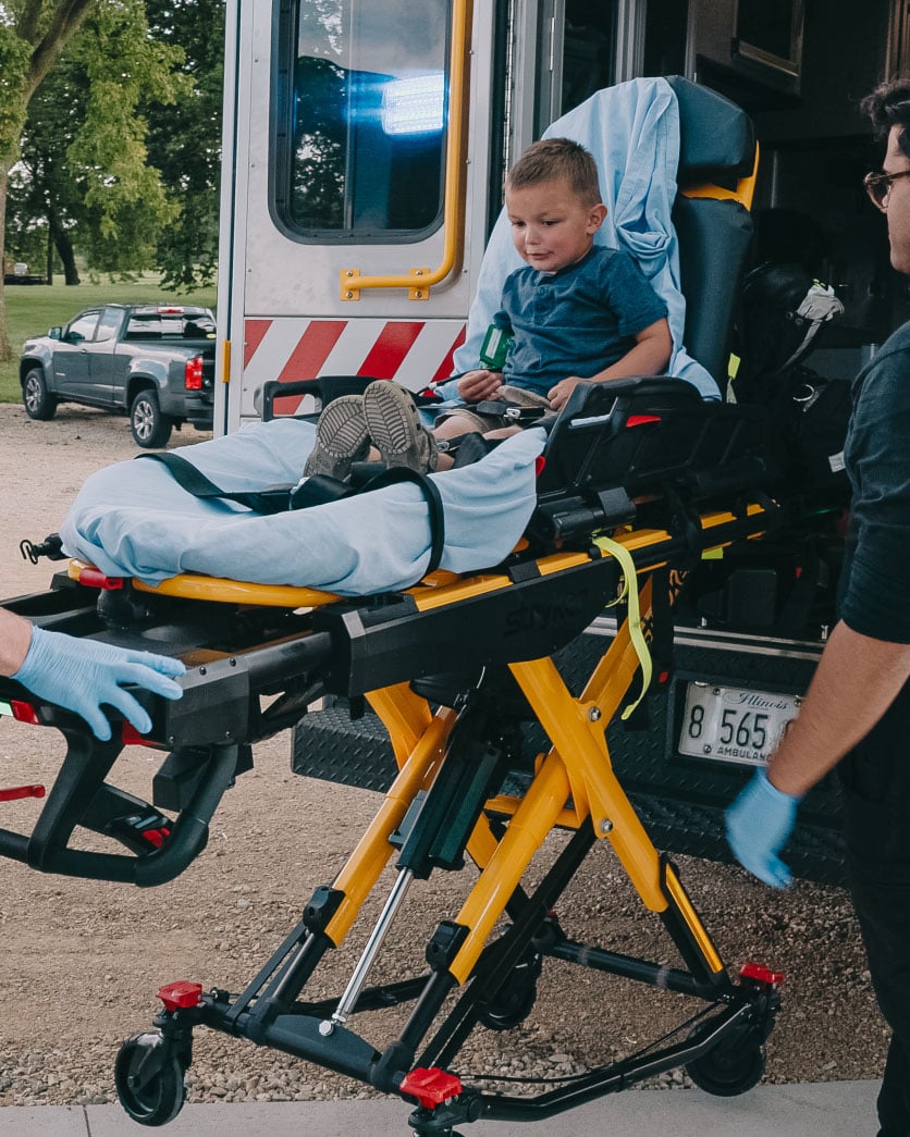 Young boy sitting on a hospital stretcher outside an ambulance with medical personnel nearby.