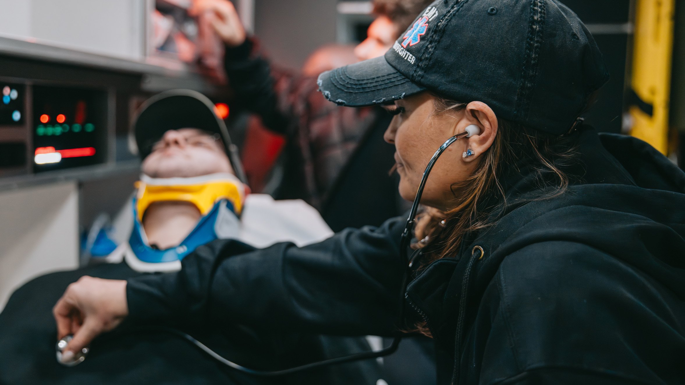 Paramedic woman wearing a black cap using a stethoscope to check a patient with a neck brace inside an ambulance.