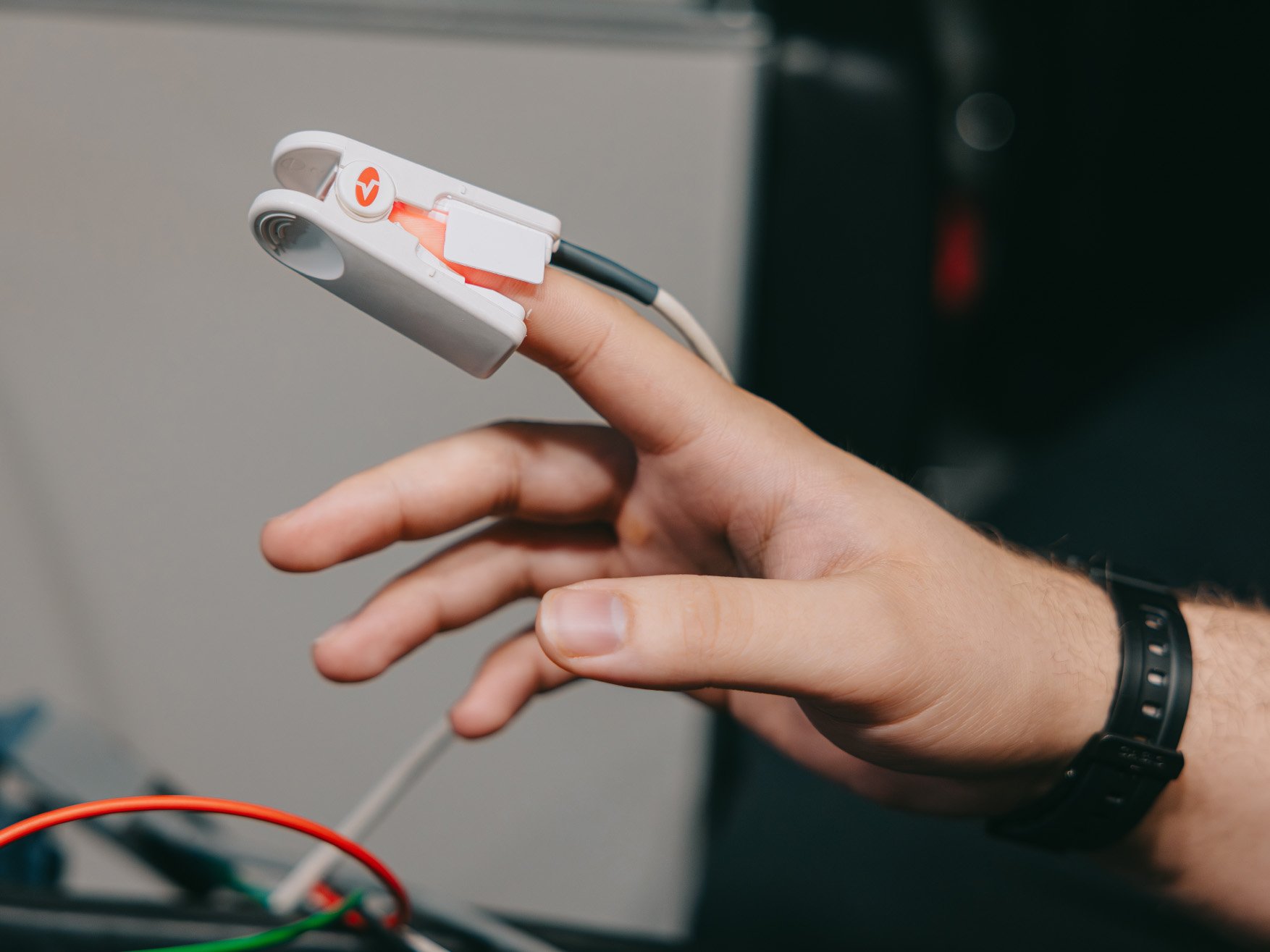 Hand with a white pulse oximeter clipped on the index finger, attached to wires.
