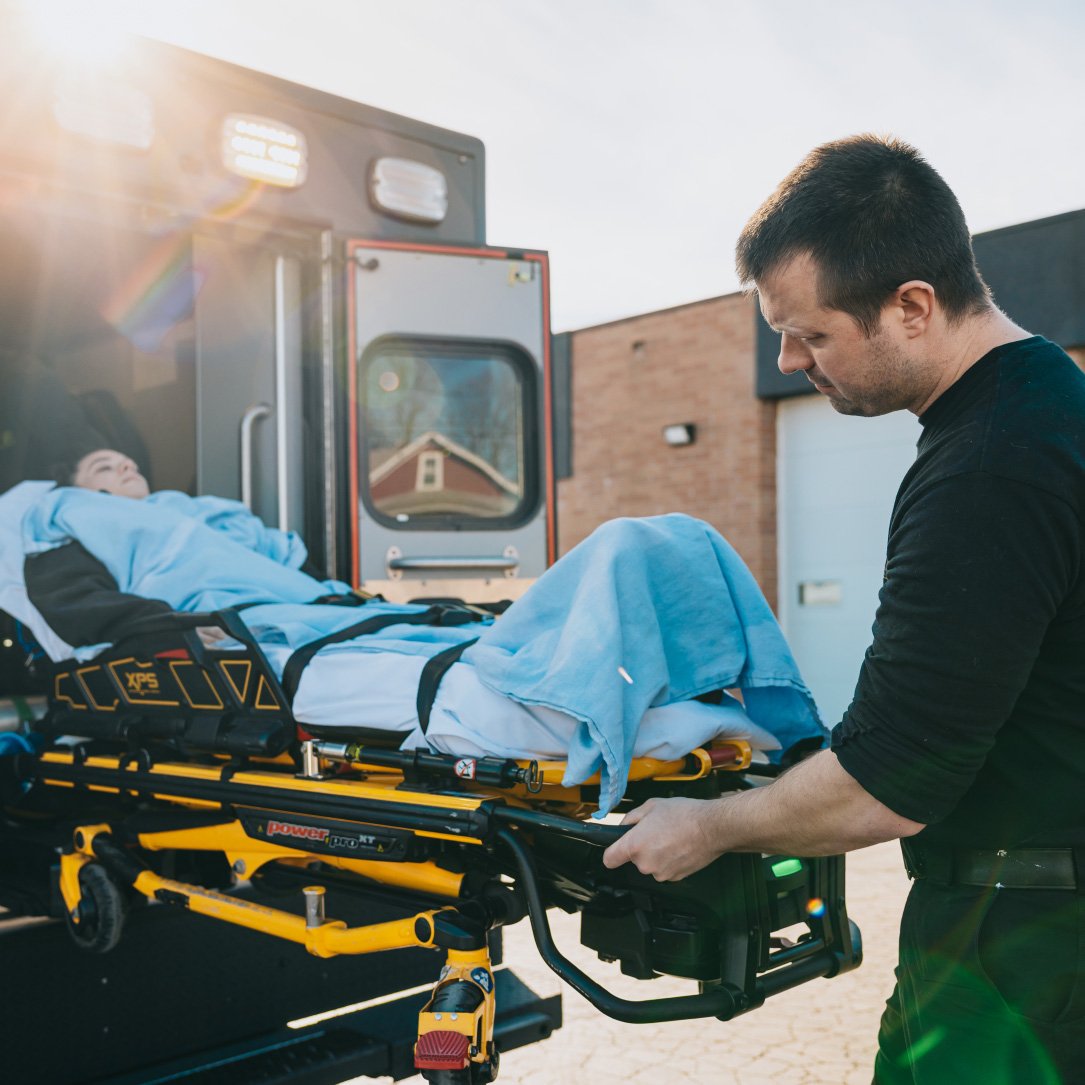 Paramedic loading a patient covered with a blue blanket into an ambulance stretcher outside a building.