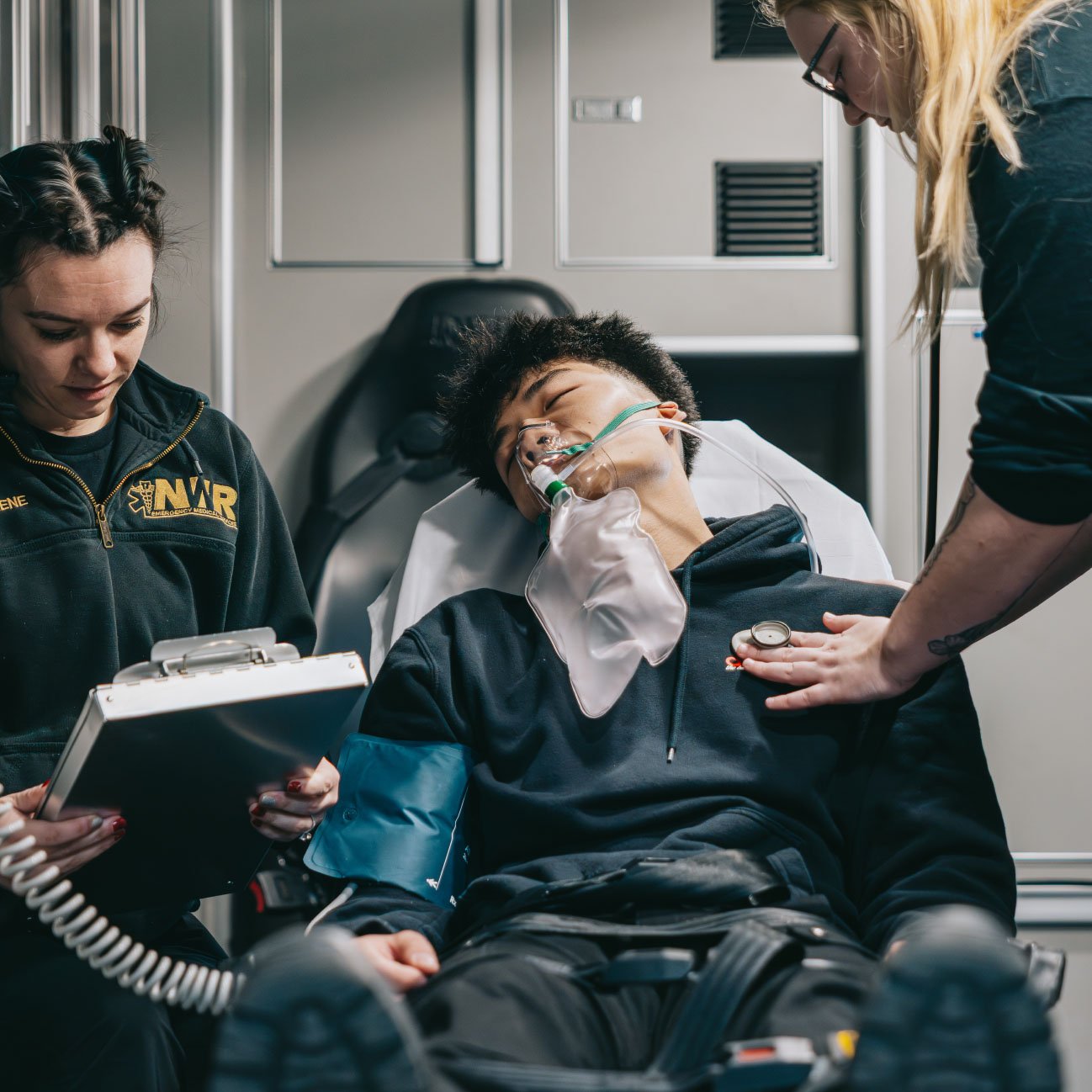 Medical responders attending to an unconscious young man on a stretcher with an oxygen mask and blood pressure cuff in an ambulance.