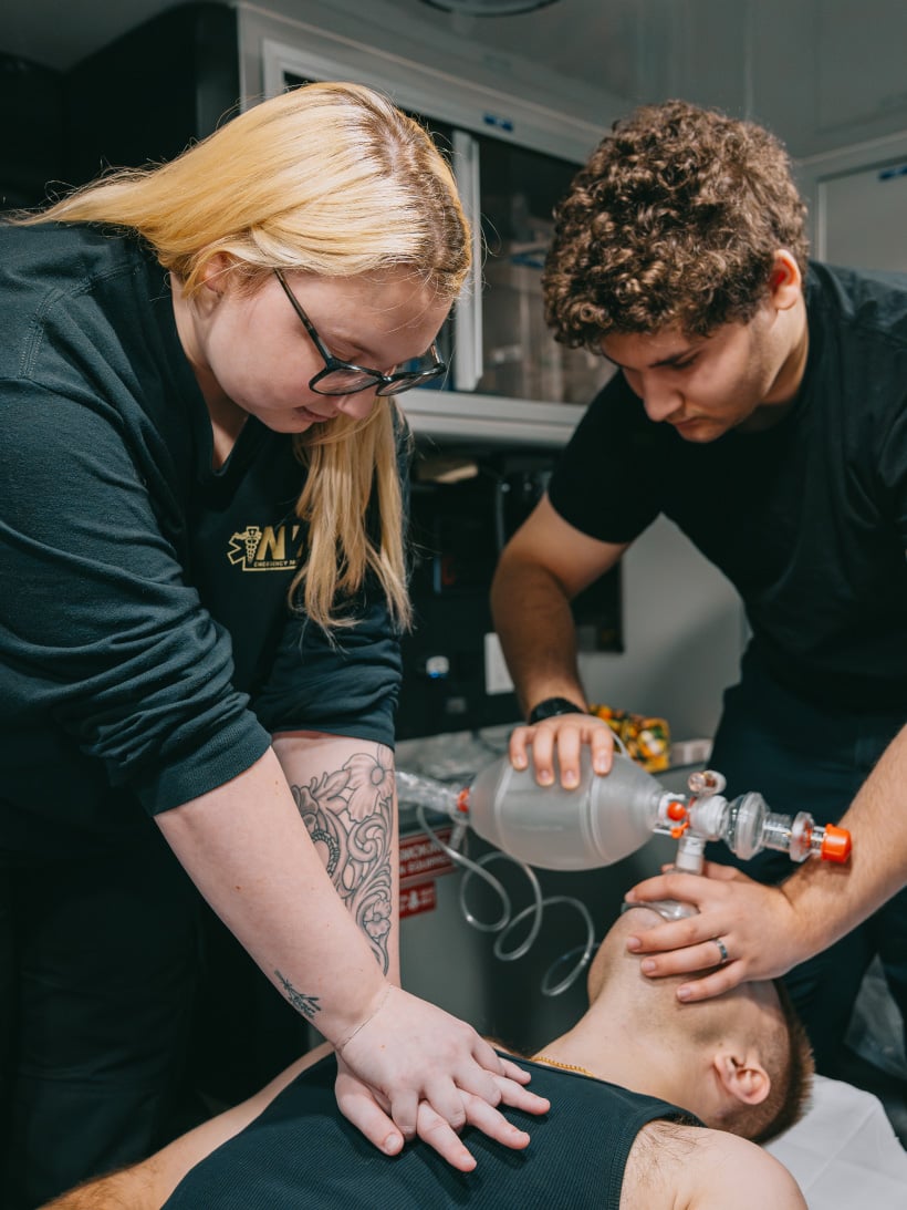 Two emergency responders performing CPR and using a bag valve mask on a person lying on a stretcher inside an ambulance.