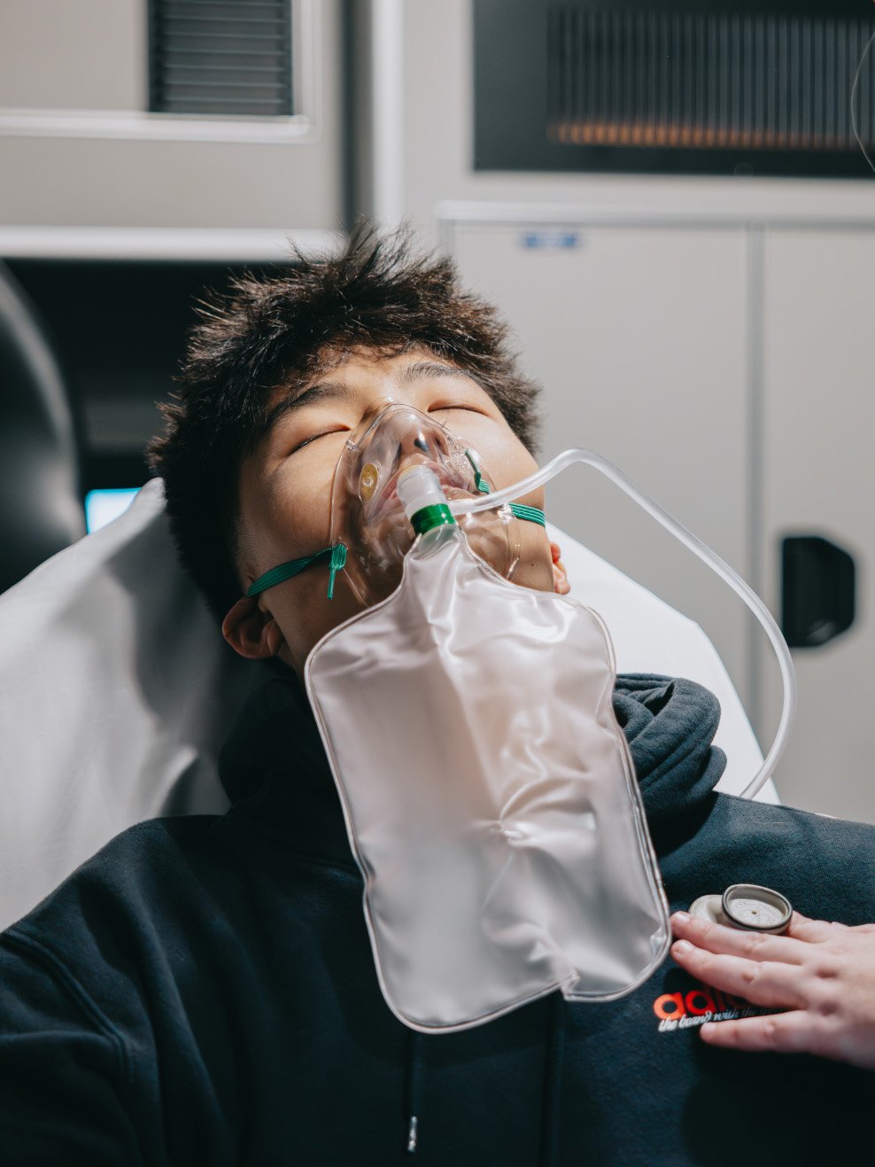 Young man lying on hospital bed wearing an oxygen mask connected to a bag, with a hand holding a stethoscope on his chest.