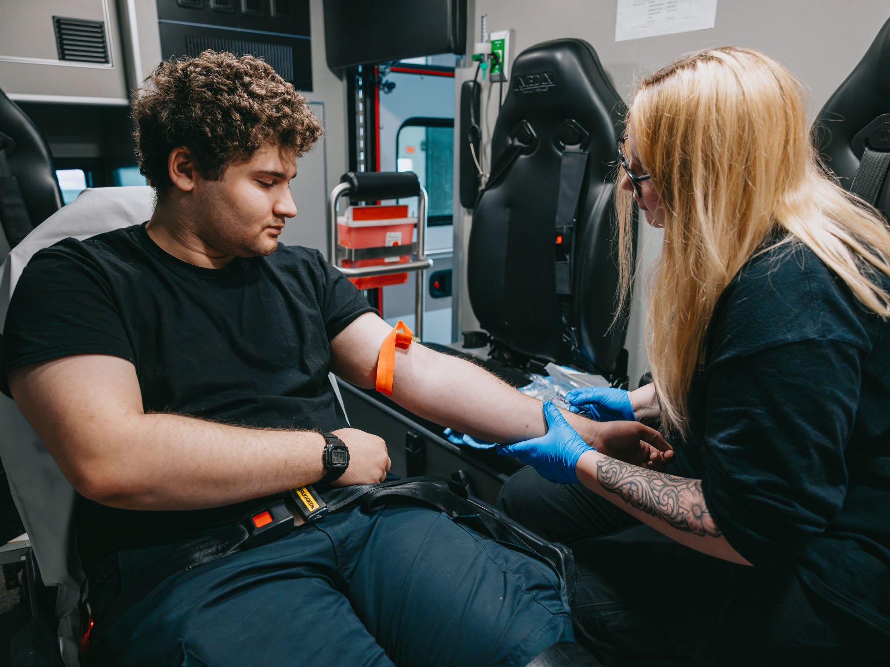 Medical professional wearing blue gloves preparing a young man’s arm for blood donation inside an ambulance.