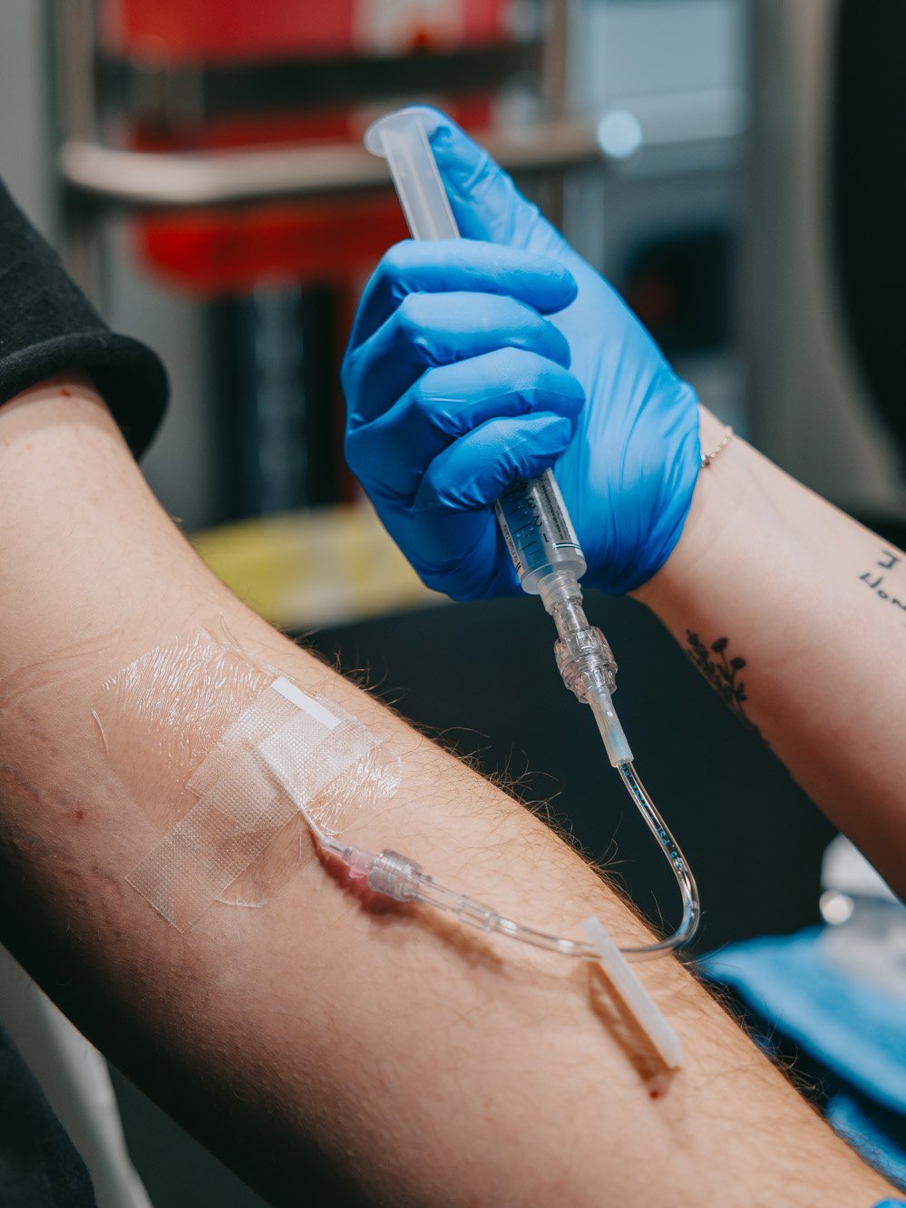 Healthcare worker wearing blue gloves holding a syringe connected to an intravenous line in a patient's arm.