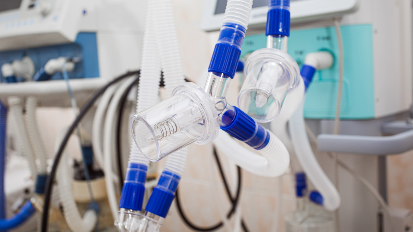 Close-up of medical ventilator tubing with clear plastic connectors and blue fittings attached to a hospital ventilator machine.