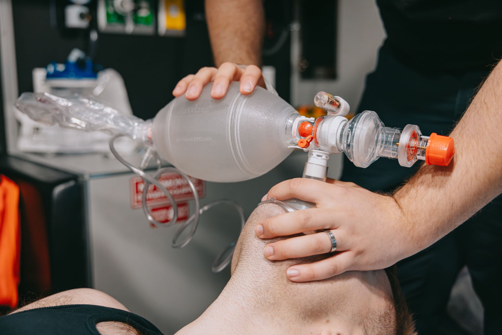 Person administering manual resuscitation using a bag valve mask on another person lying down.