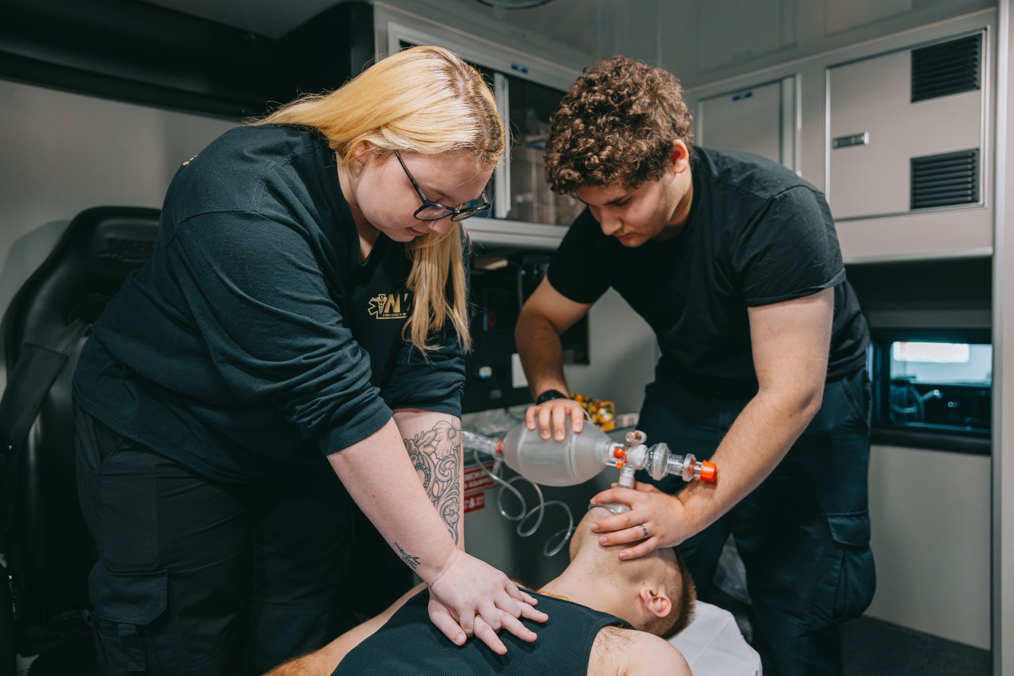 Two paramedics providing CPR and using a manual resuscitator on a patient lying on a stretcher inside an ambulance.