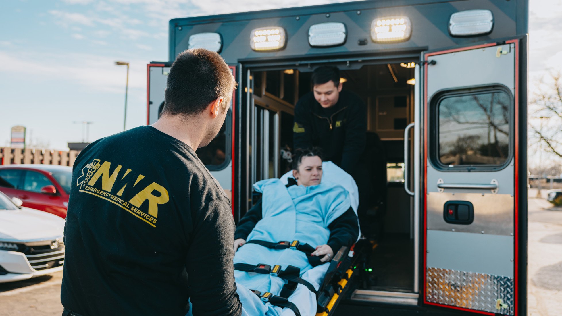 Emergency medical responders loading a woman wrapped in a blue blanket onto a stretcher into an ambulance.
