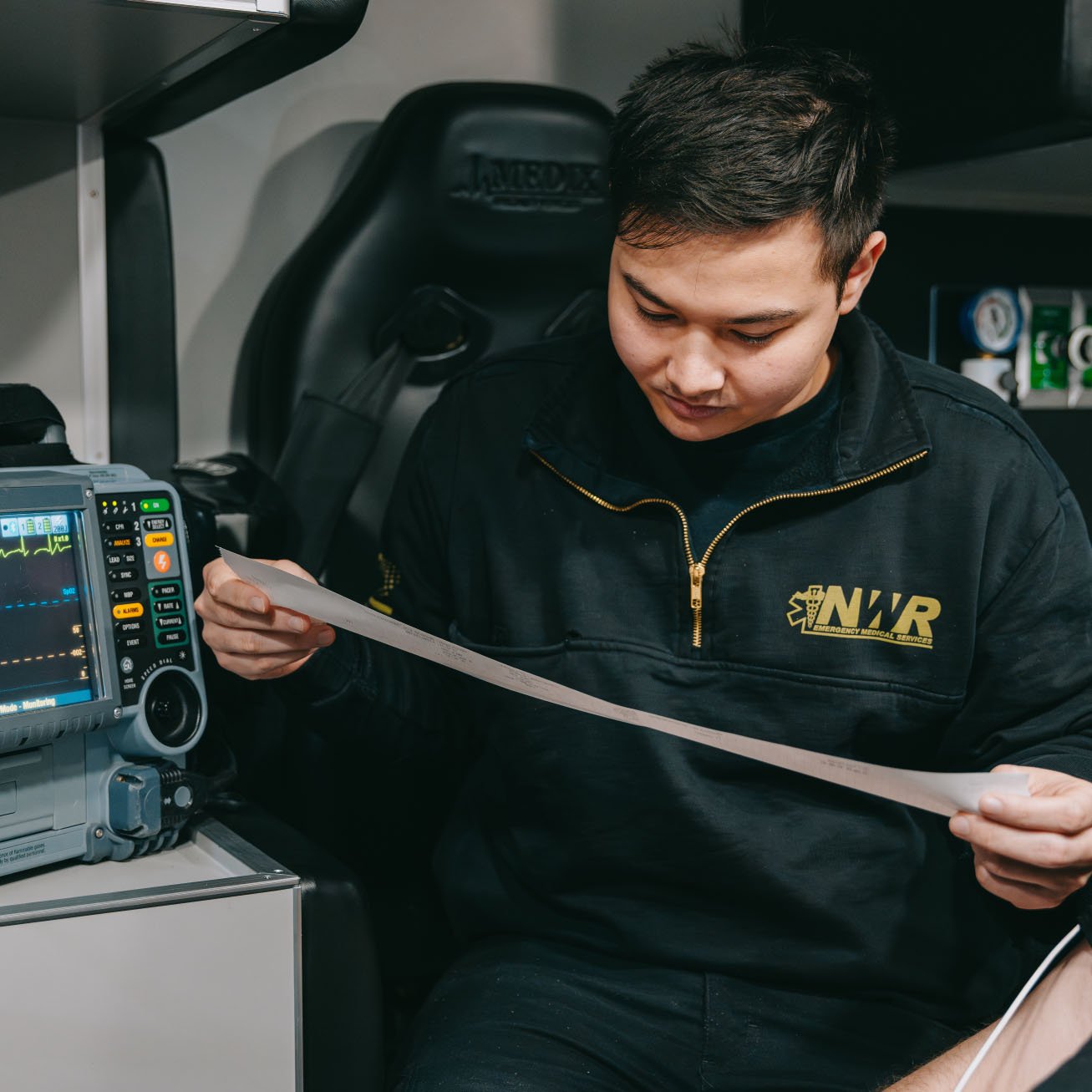 Emergency medical technician in a black NWR jacket reviewing a long medical printout inside an ambulance.