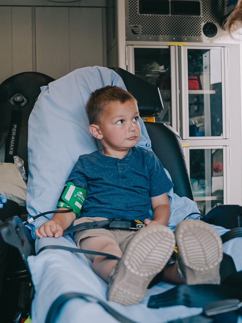 Young boy sitting on a stretcher inside an ambulance, wearing a blue shirt and a blood pressure cuff on his arm.