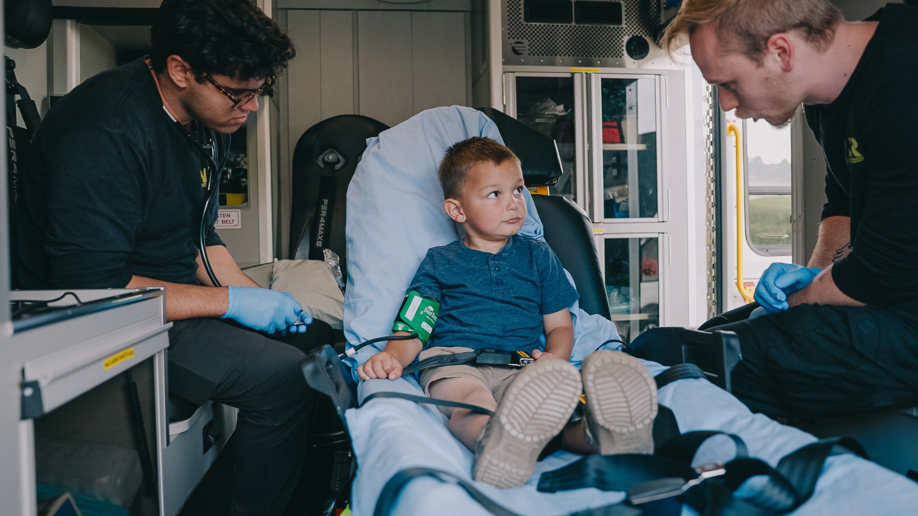 Young boy sitting on a stretcher inside an ambulance with medical personnel wearing gloves attending to him.