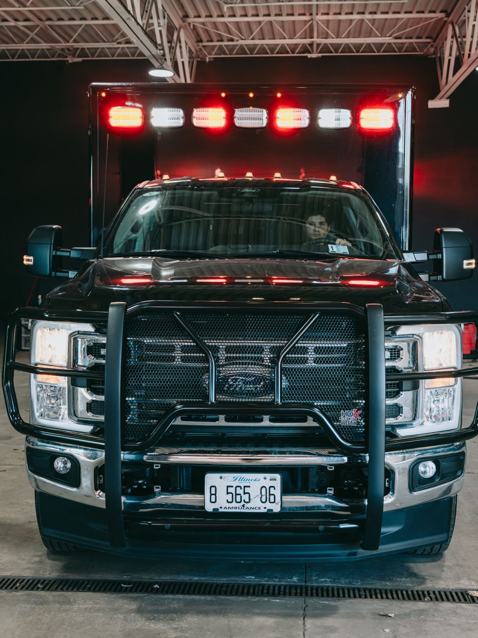 Front view of a black ambulance with red and white emergency lights on the roof inside a garage.
