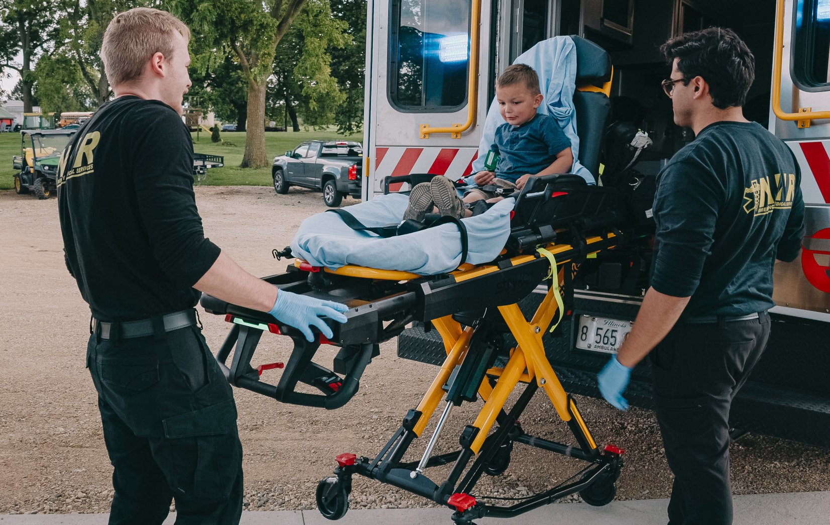 Two emergency medical responders loading a smiling young boy on a stretcher into an ambulance.
