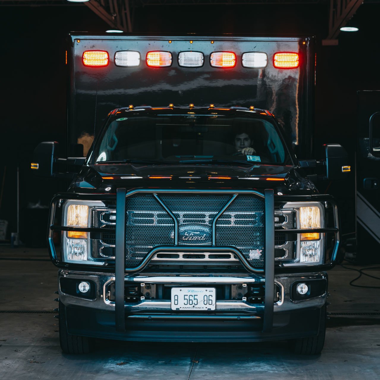 Front view of a black Ford ambulance with lights on and a person visible behind the steering wheel in a dimly lit garage.