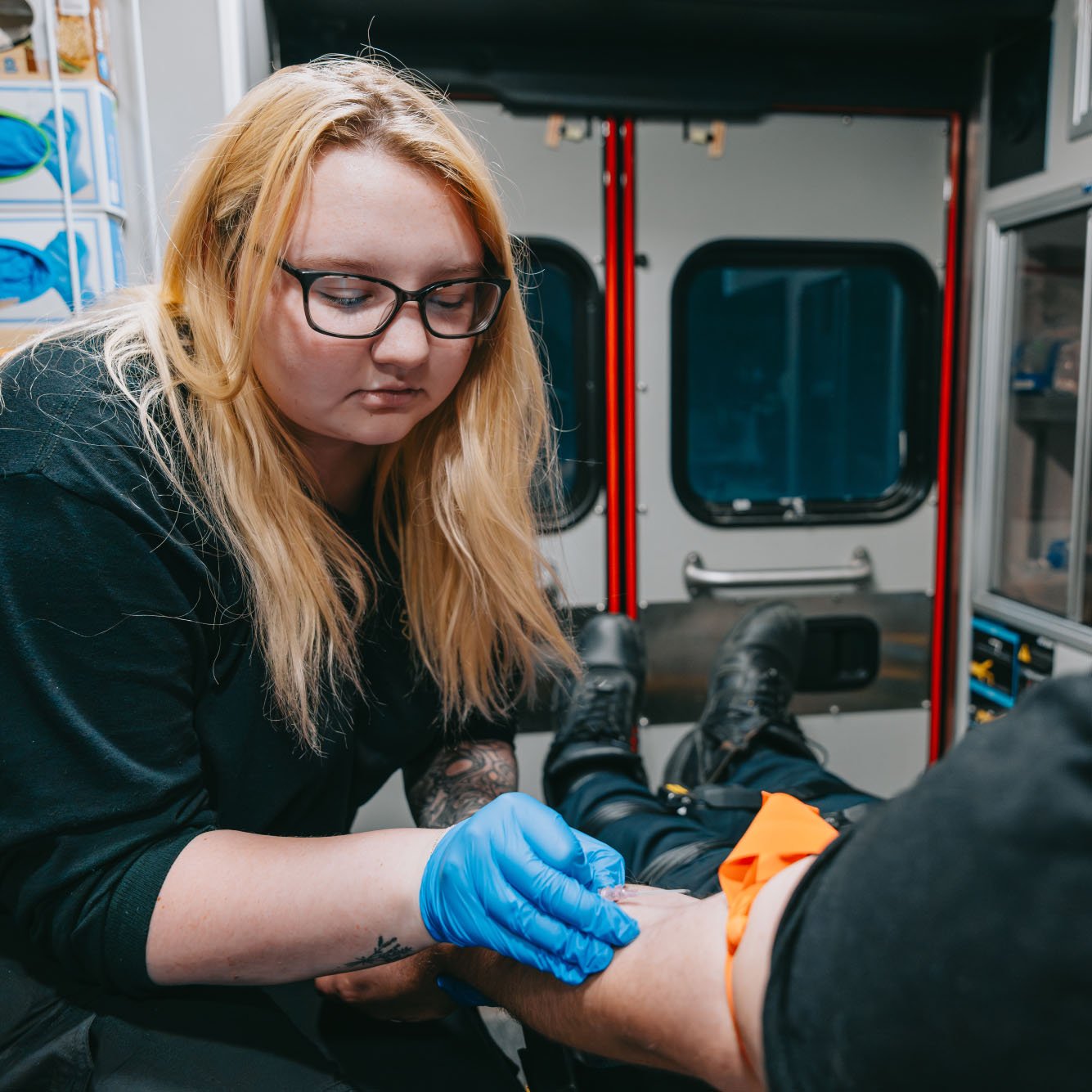 Paramedic with blonde hair and glasses wearing blue gloves attending to a patient's arm inside an ambulance.
