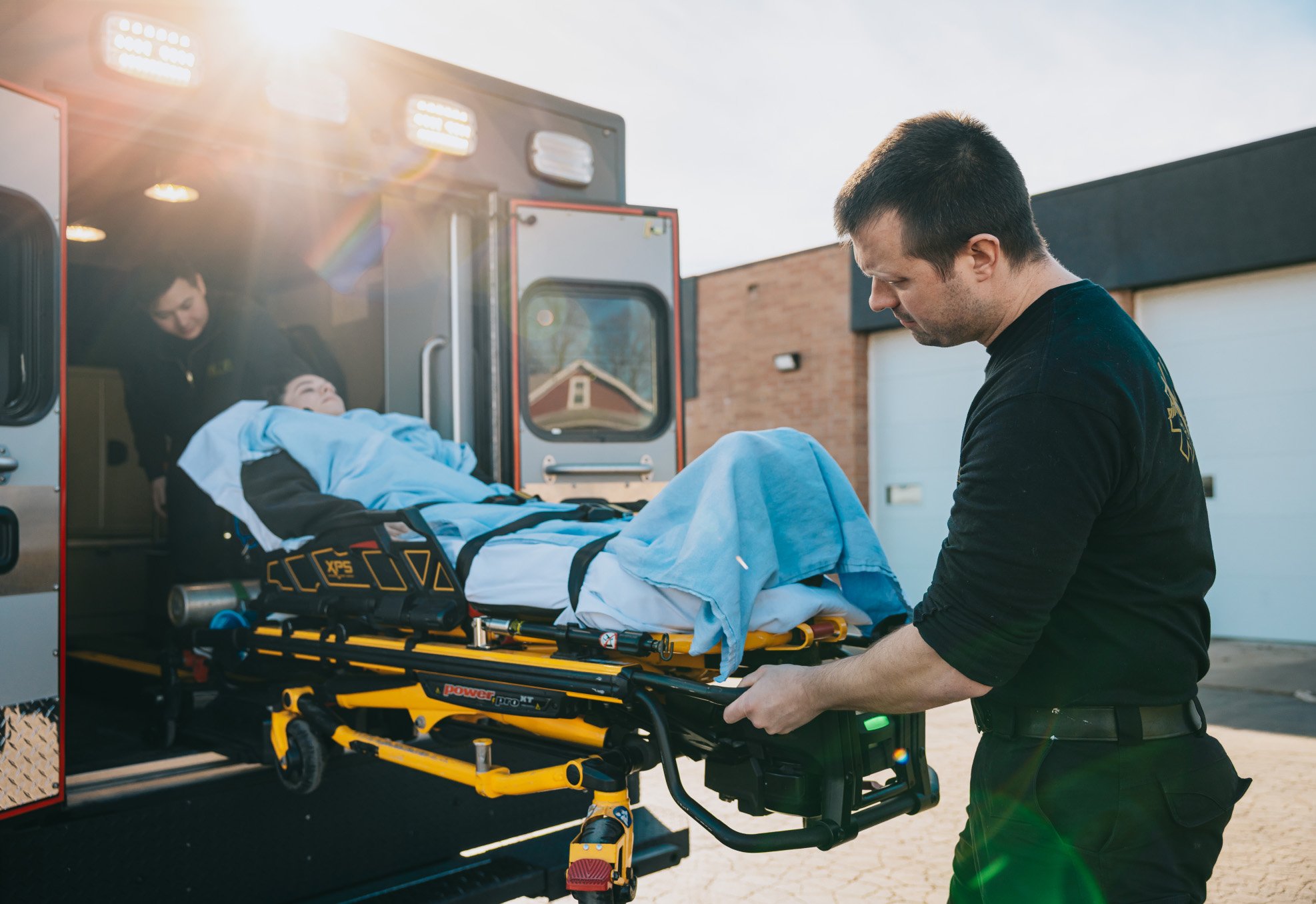 Paramedics loading a patient covered with a blue blanket on a stretcher into an ambulance.