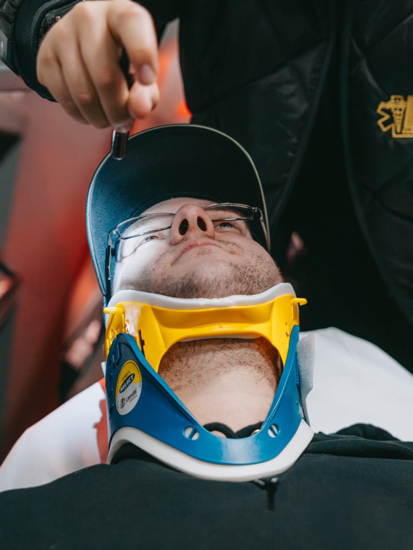 Close-up of a person wearing a blue and yellow neck brace and glasses, being assisted by another person holding a small object above them.