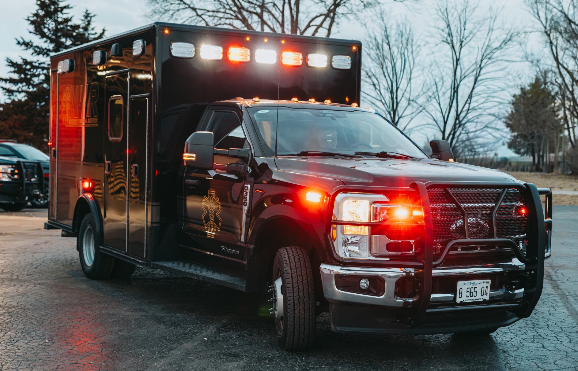 Black ambulance with flashing emergency lights parked on a cracked pavement at dusk.