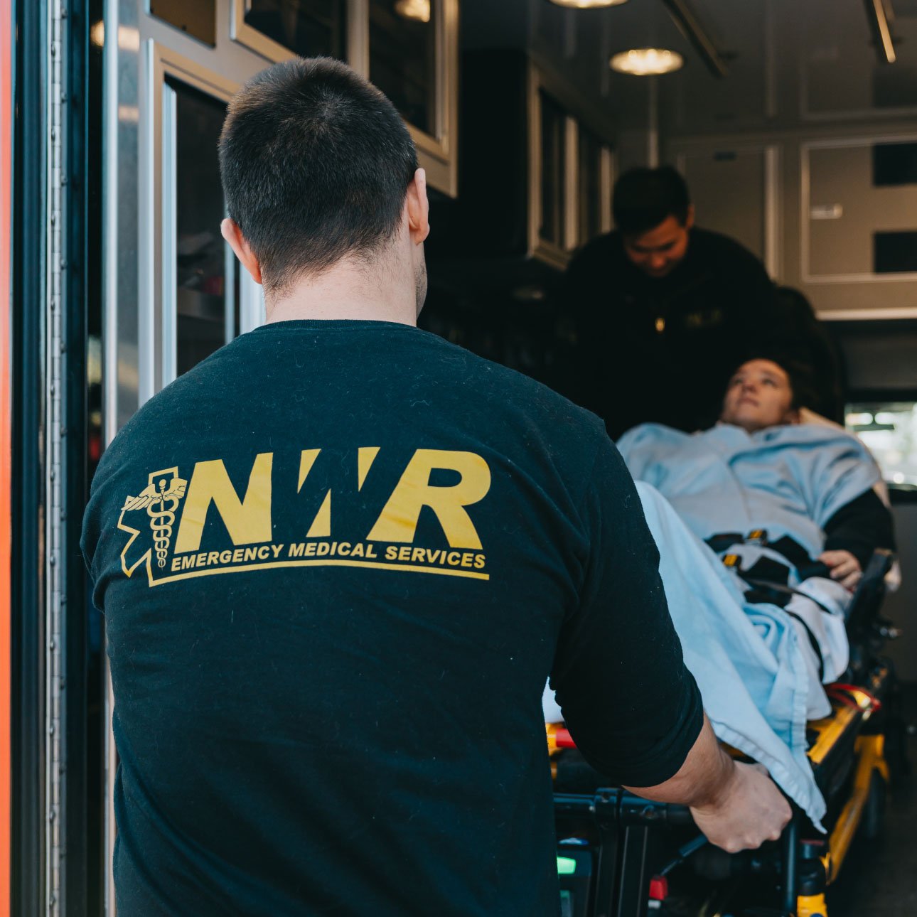 Paramedic wearing a black NWR Emergency Medical Services shirt pushing a patient on a stretcher inside an ambulance.