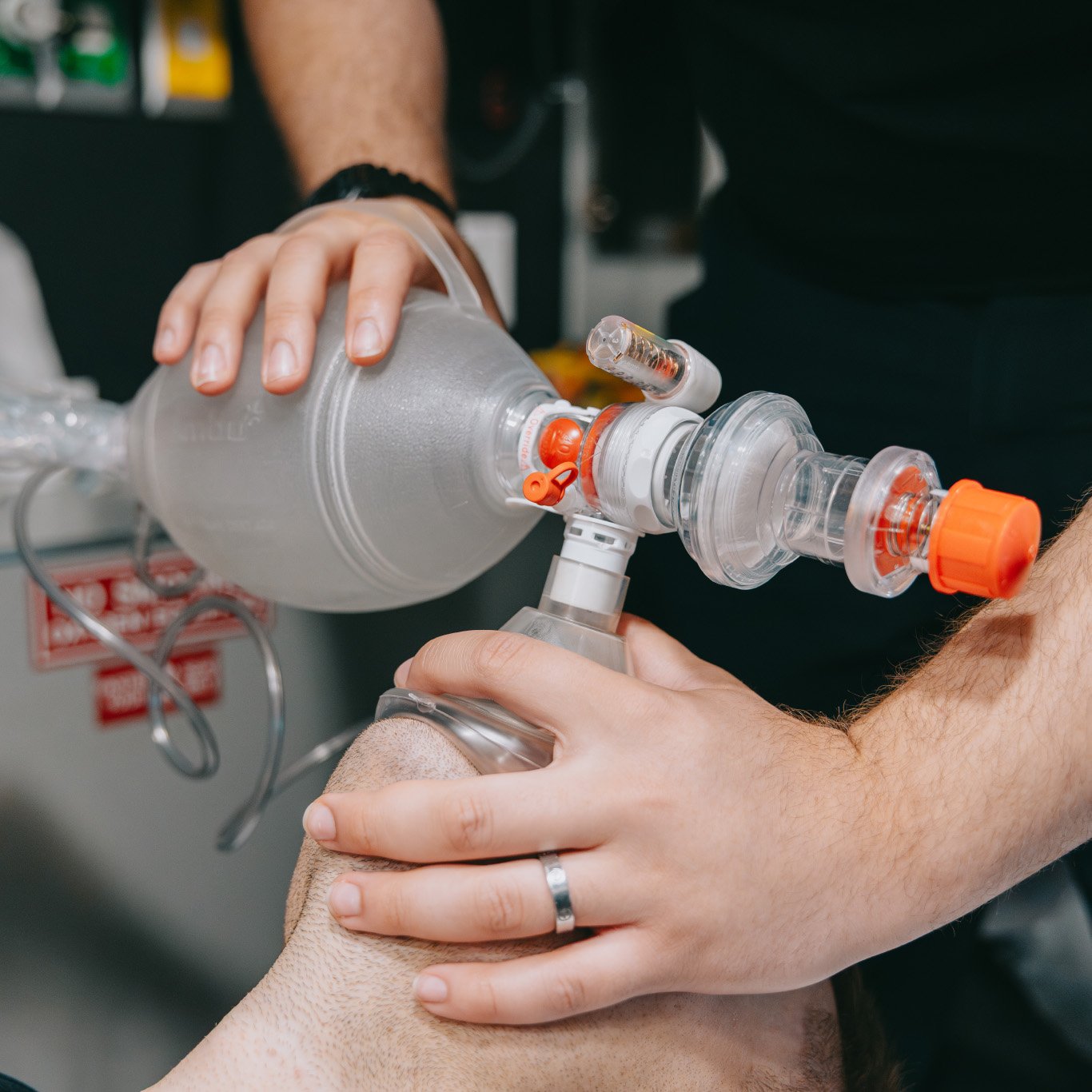 Close-up of hands using a manual resuscitator to provide ventilation to a person wearing an oxygen mask.