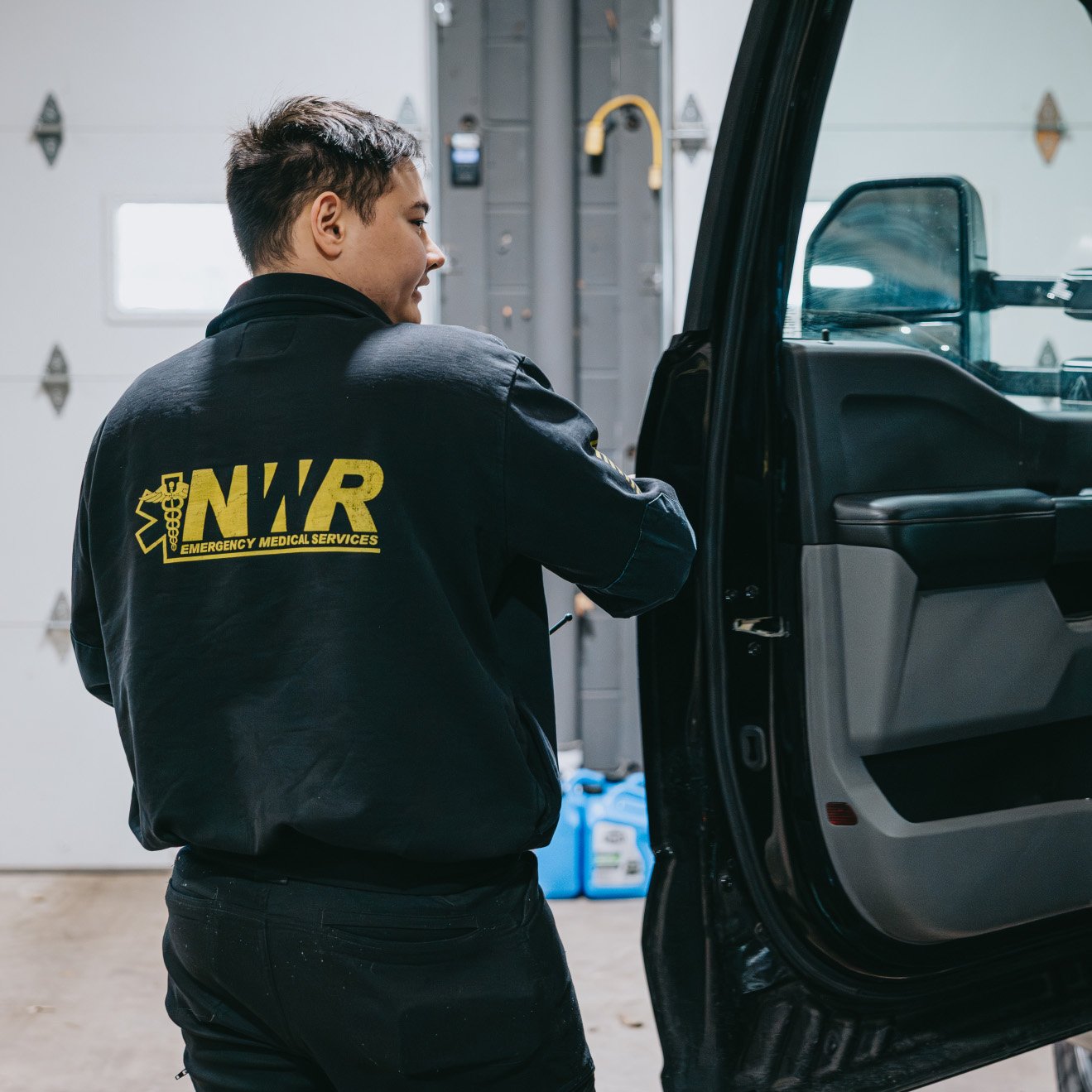 Person wearing a black jacket with yellow NWR Emergency Medical Services logo opening a black vehicle door in a garage.