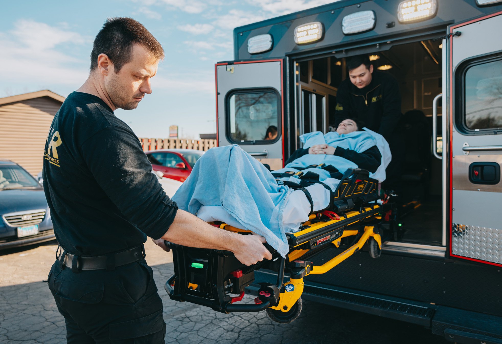 Paramedics loading a patient covered with a blue blanket on a stretcher into an ambulance.