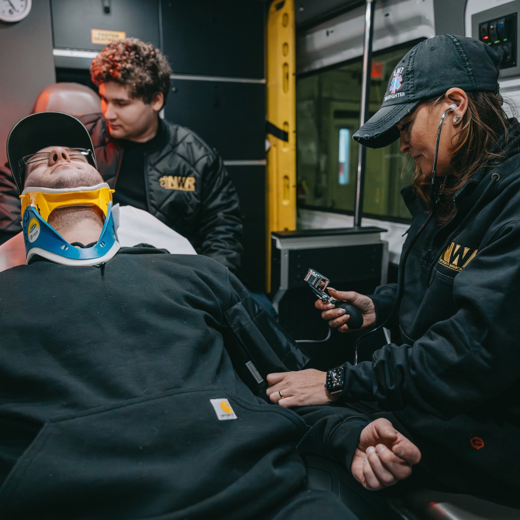EMT checking blood pressure of a patient wearing a neck brace inside an ambulance, with another EMT observing in the background.