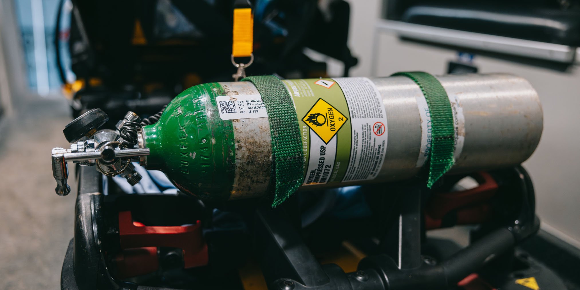 Compressed oxygen gas cylinder with green top and hazard warning label secured by green straps on equipment.