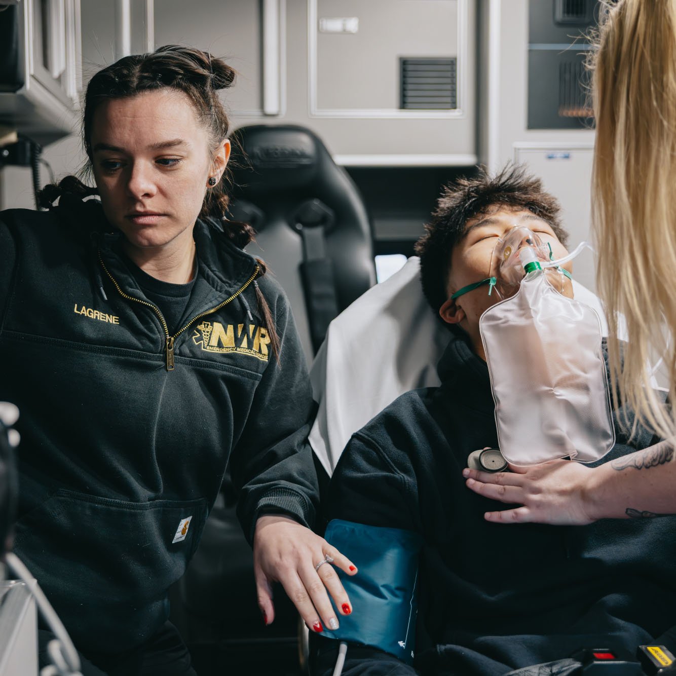 Emergency medical technician attending to a patient with an oxygen mask and blood pressure cuff inside an ambulance.