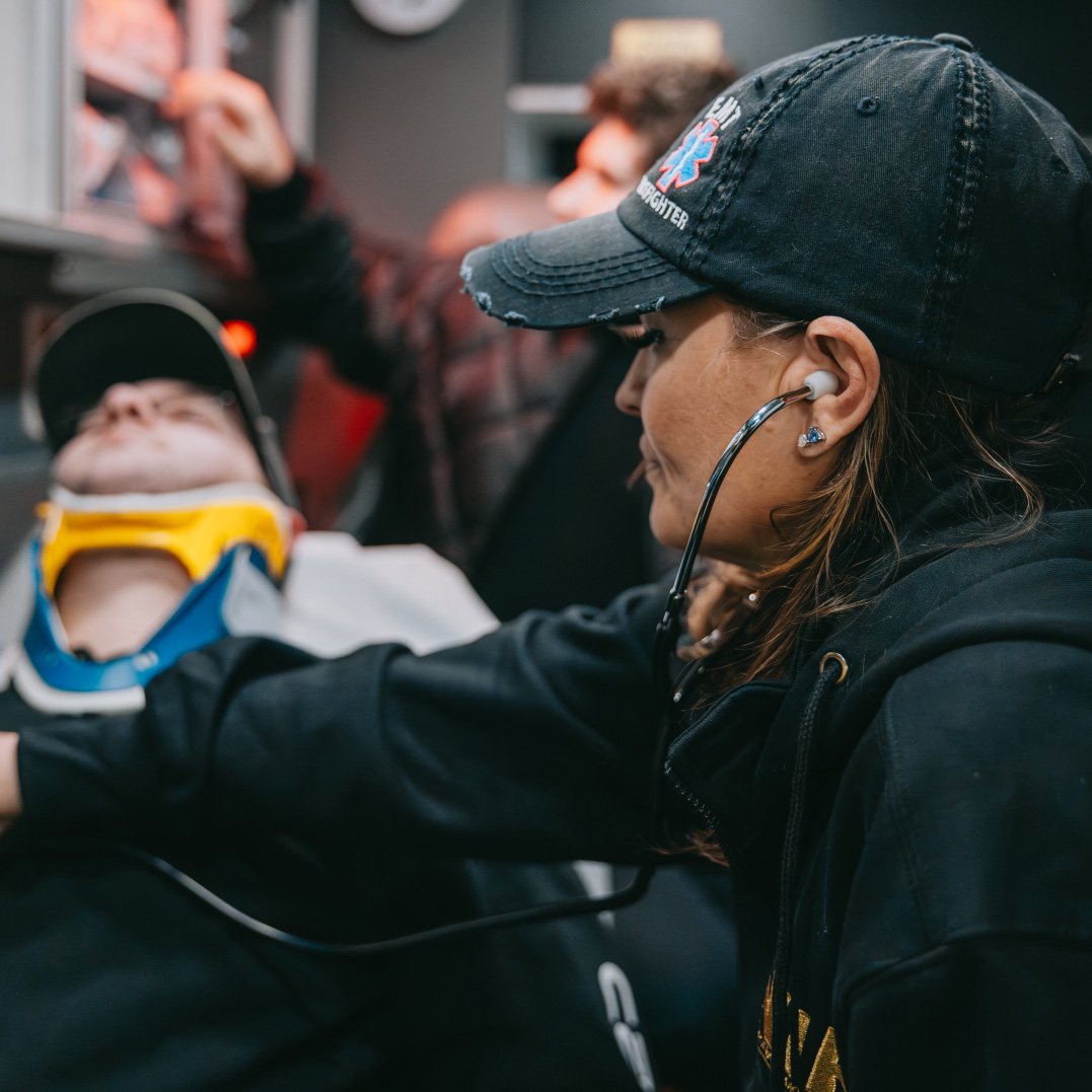 Emergency responder wearing a black cap uses a stethoscope to check a patient with a neck brace lying on a stretcher.
