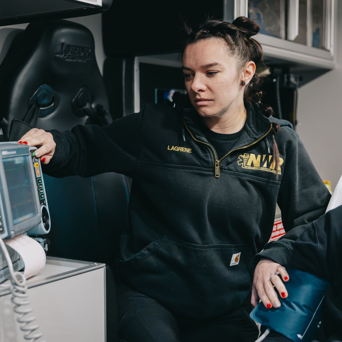 Female paramedic in black NW paramedic sweatshirt checking medical monitor inside an ambulance.