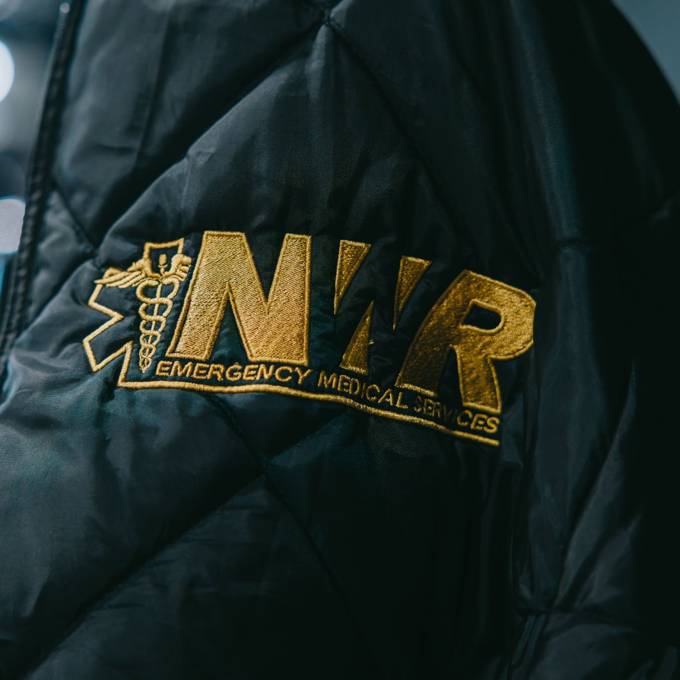 Close-up of black quilted fabric with gold embroidered letters 'NWR Emergency Medical Services' and a medical caduceus symbol.
