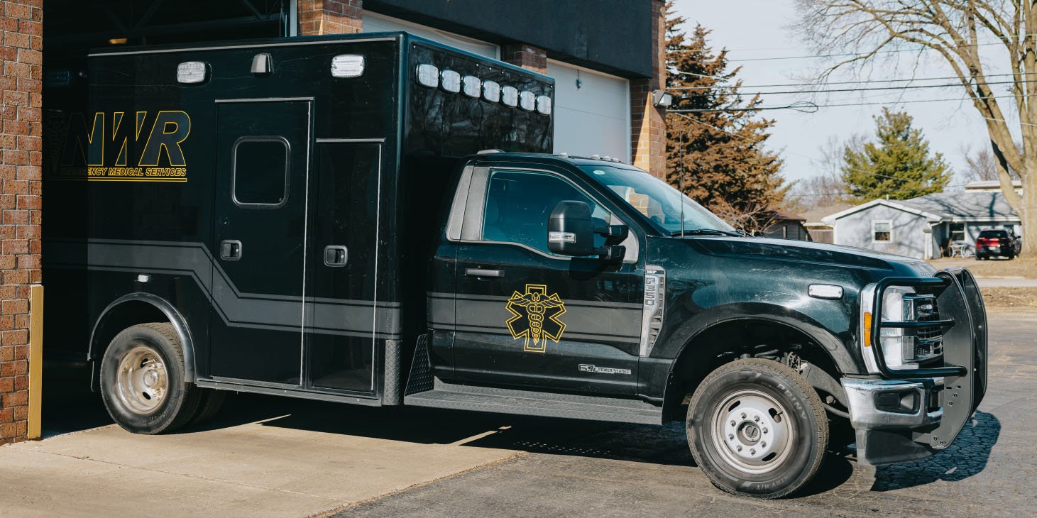 Black emergency medical services ambulance truck parked partially inside a brick garage.