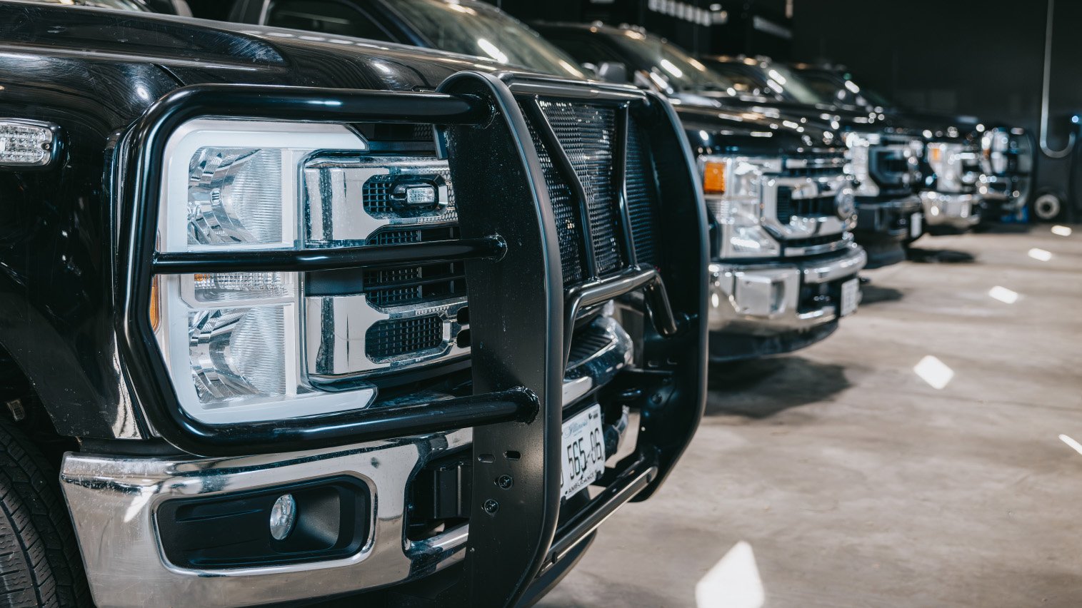 Close-up of the front end of black pickup trucks with grille guards lined up indoors on a concrete floor.