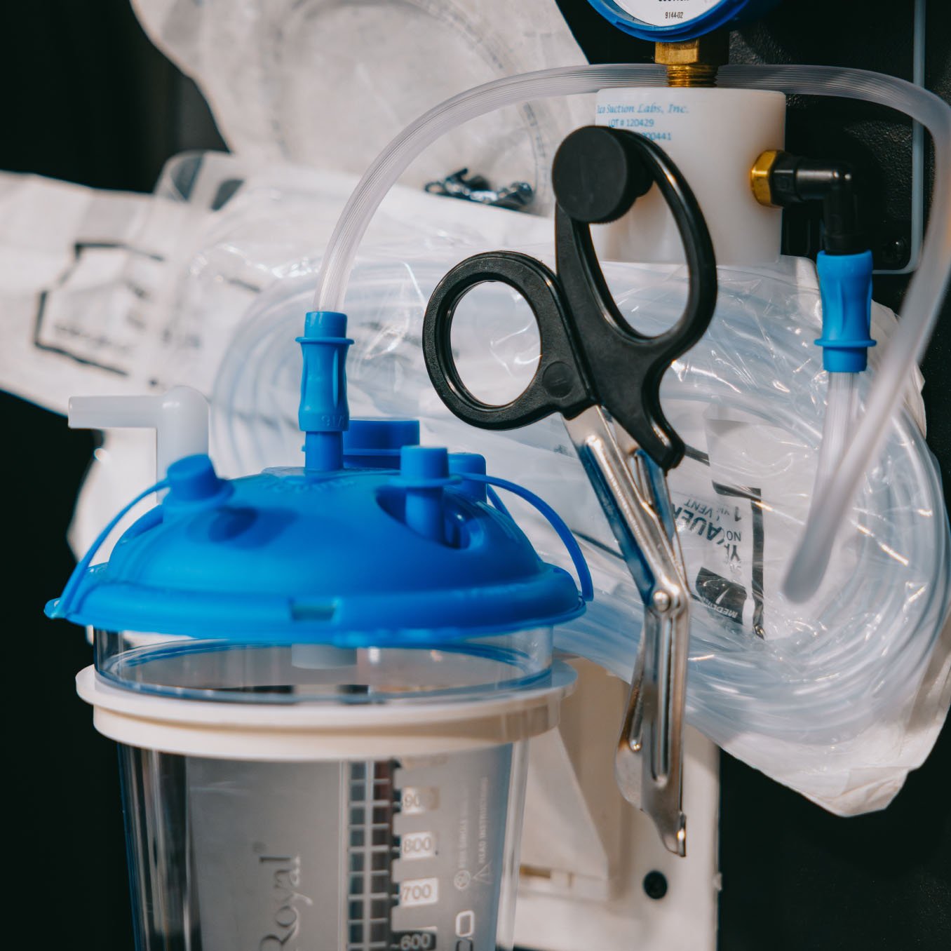Close-up of black-handled medical scissors clipped to a transparent container with a blue lid and connected tubes.