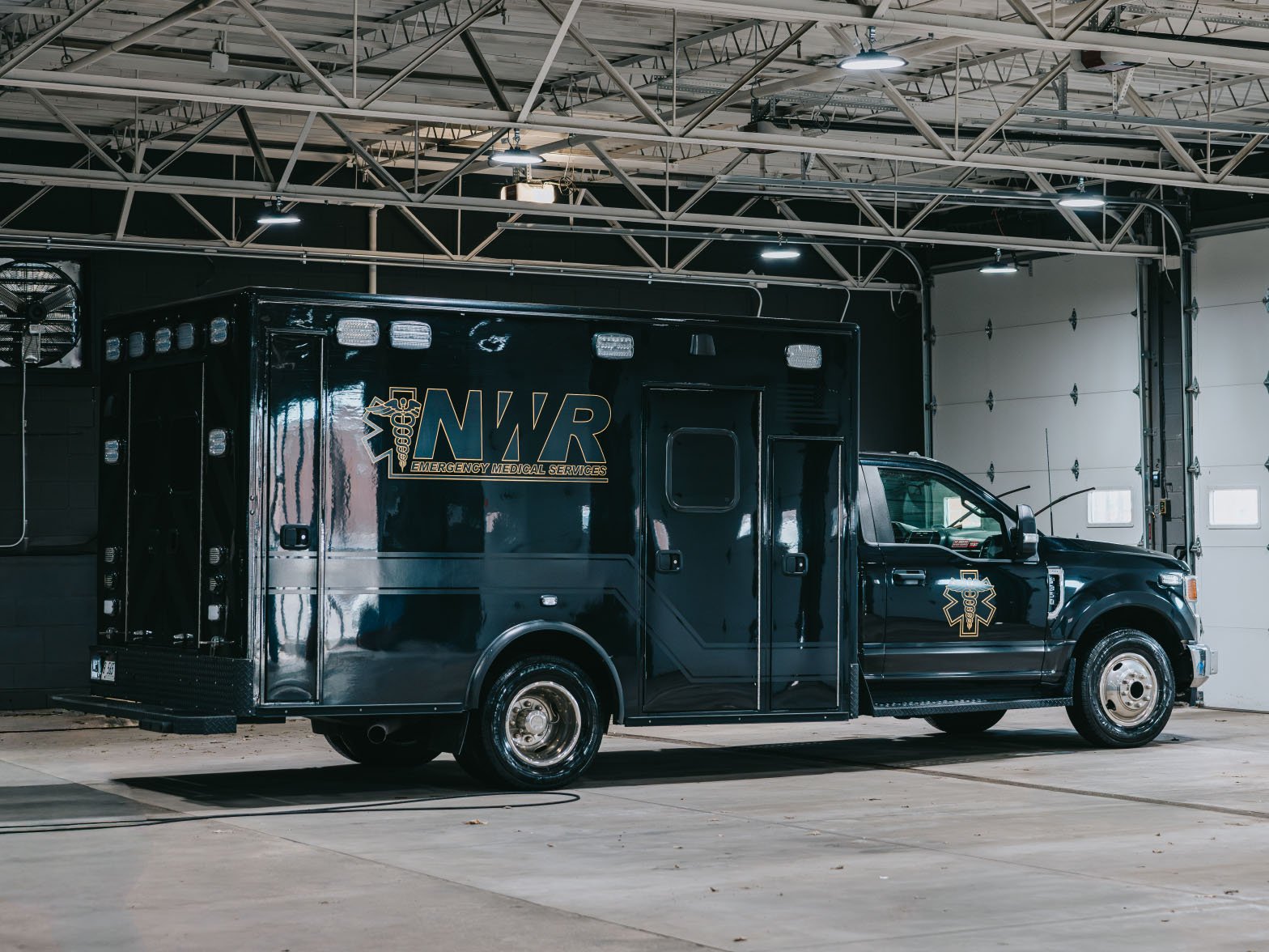 Black emergency medical services truck with NWR logo parked inside a garage with high ceiling and industrial lights.