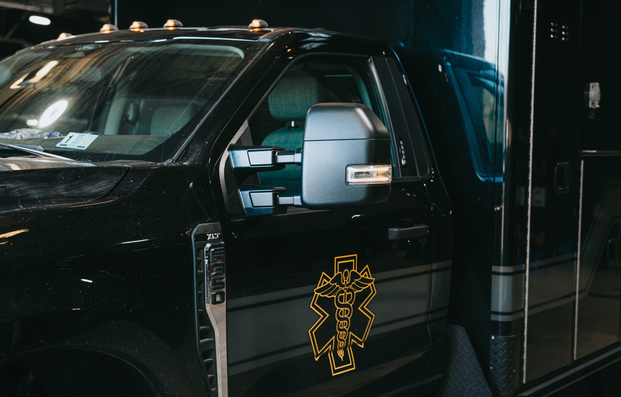 Side view of a black medical emergency vehicle with a gold Star of Life symbol on the door and an extended side mirror.