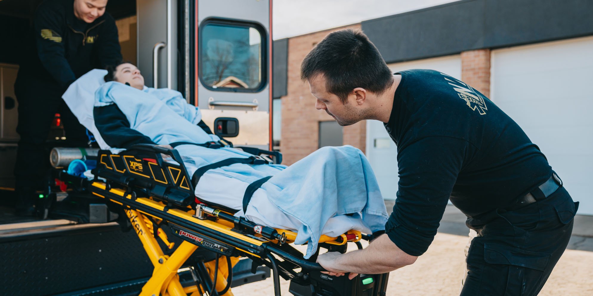 Two paramedics loading a patient covered in a light blue blanket into an ambulance on a stretcher.