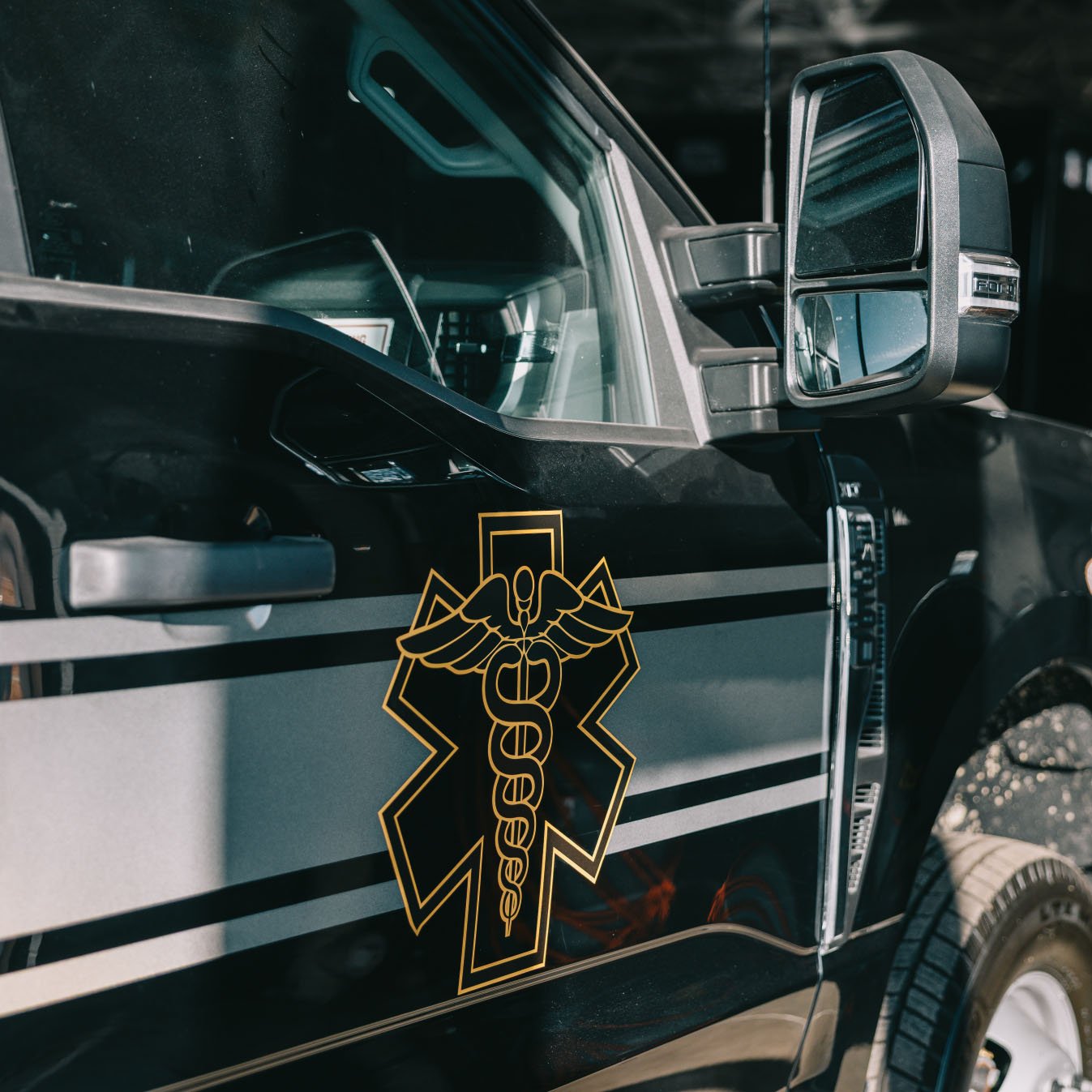 Side view of a black emergency vehicle door showing a gold Star of Life medical emblem and part of the side mirror and tire.