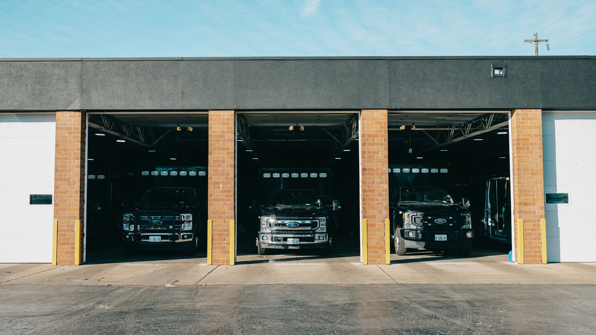 Three black Ford pickup trucks parked inside open garage bays of a brick and gray commercial building under a blue sky.