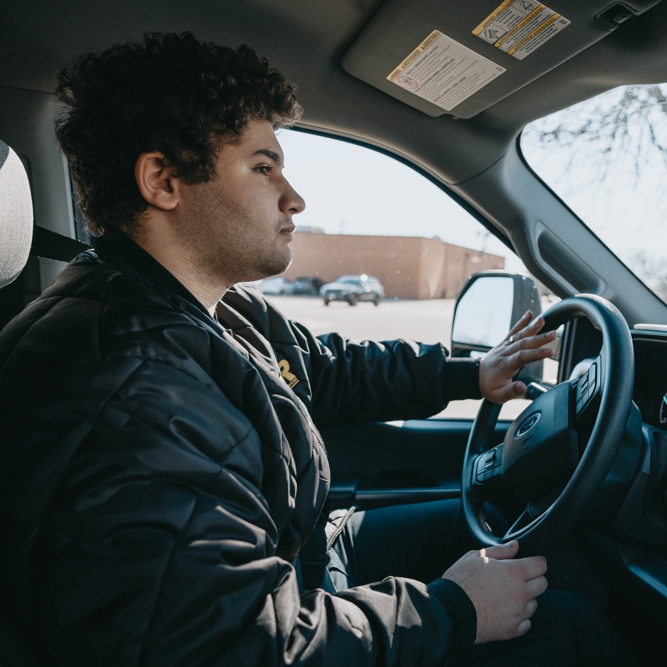 Man with curly hair wearing a black jacket, driving a car and looking forward.